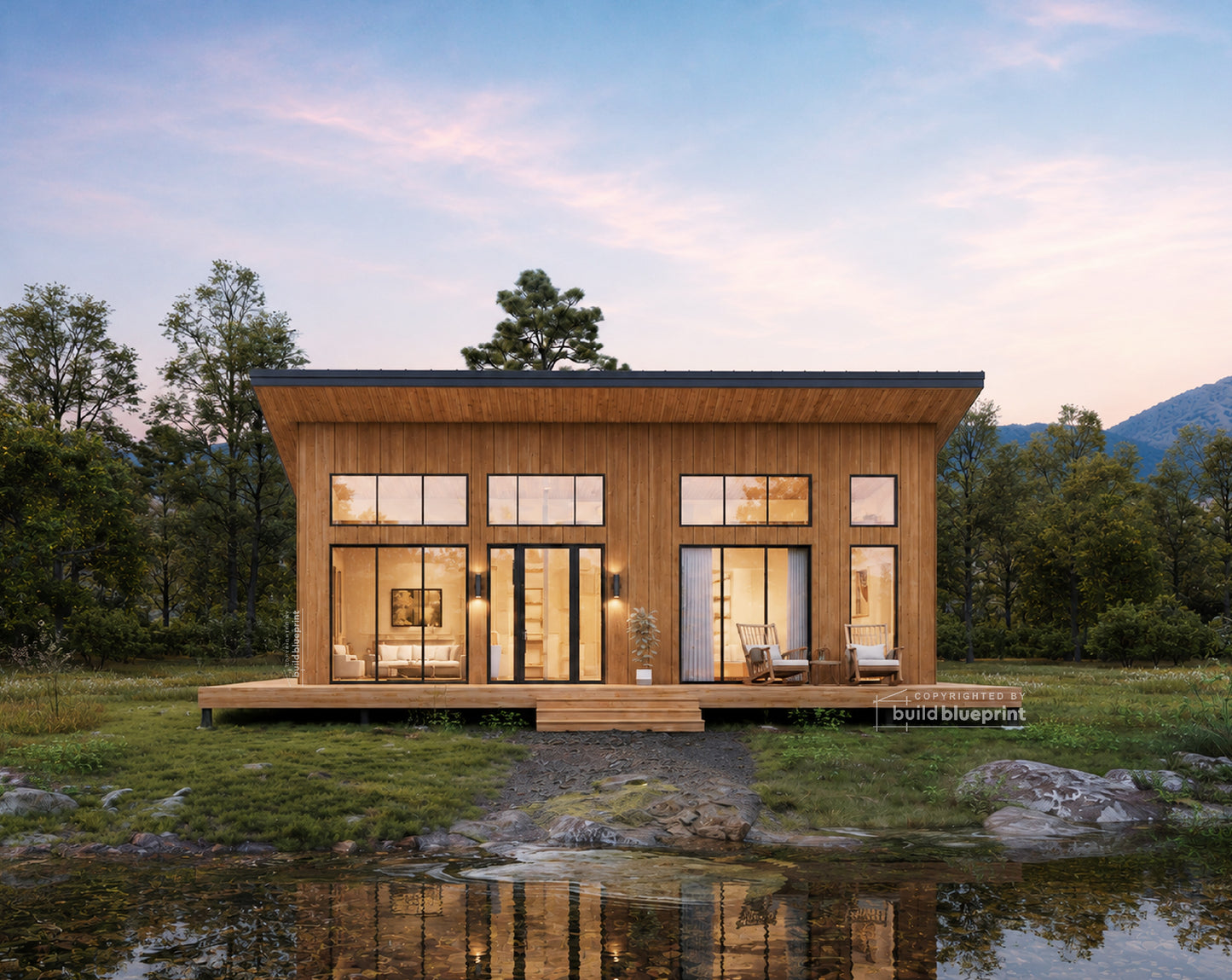 A modern single-story cabin with warm wood siding and large glass windows, situated on a grassy bank beside a reflective pond with trees and mountains in the background at dusk.