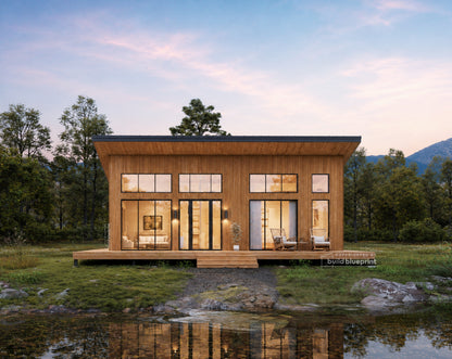 A modern single-story cabin with warm wood siding and large glass windows, situated on a grassy bank beside a reflective pond with trees and mountains in the background at dusk.