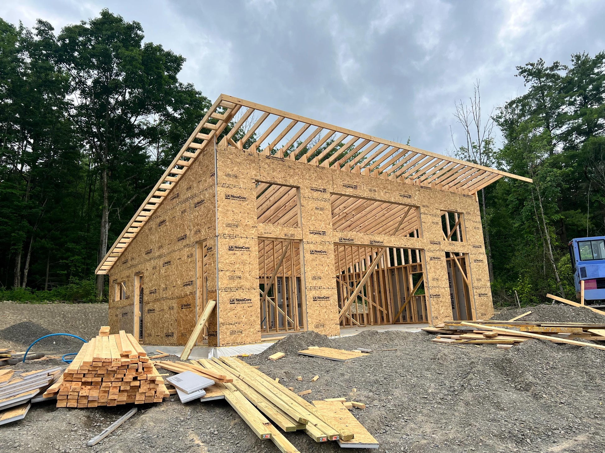 A large wooden house frame under construction, made of OSX plywood panels, with exposed roof trusses and surrounded by construction materials and trees.