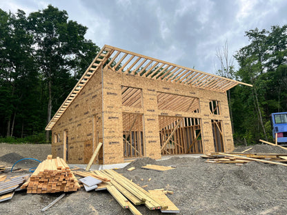 A large wooden house frame under construction, made of OSX plywood panels, with exposed roof trusses and surrounded by construction materials and trees.
