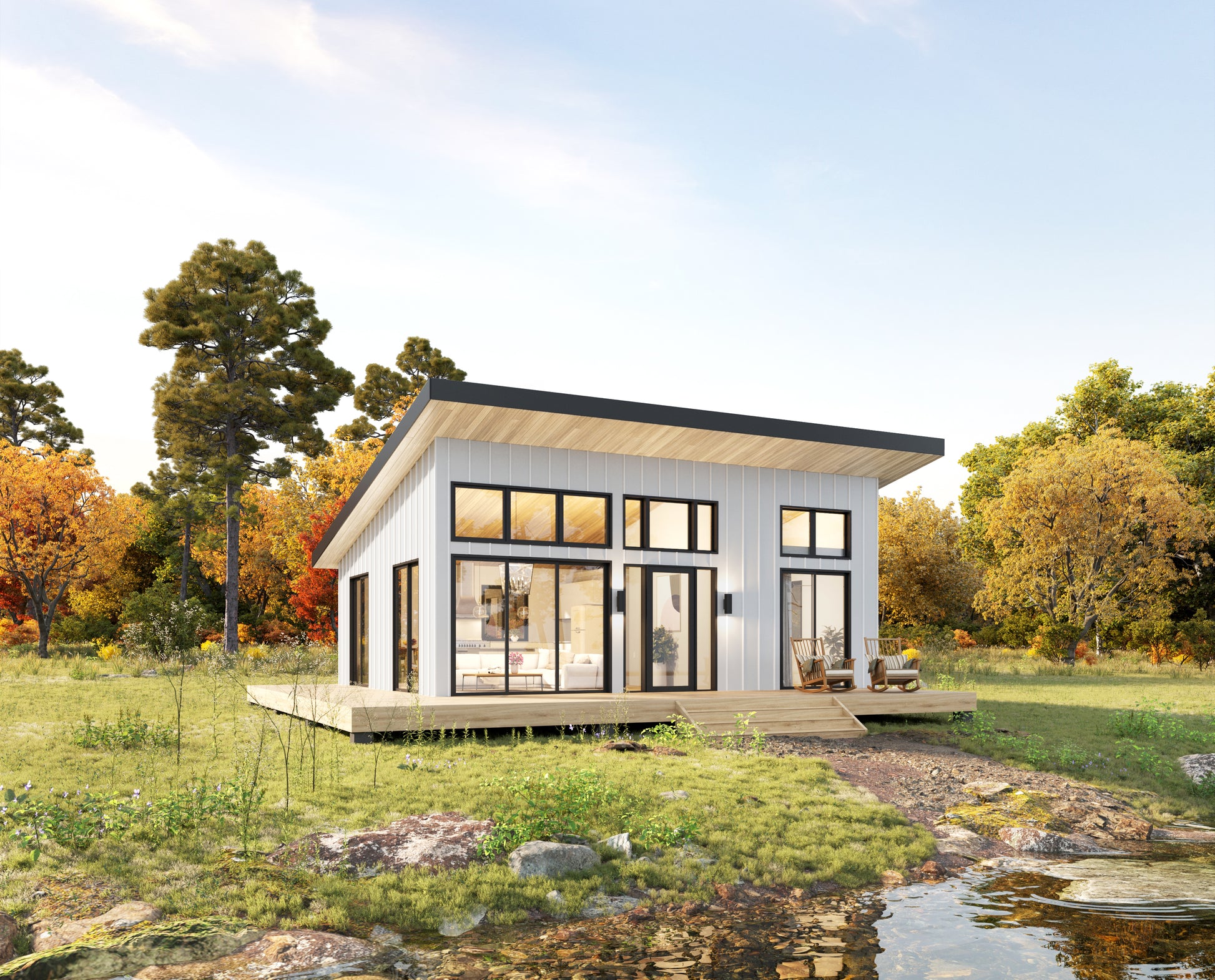 A modern single-story cabin with white siding, large black-framed windows, and a wooden deck, shown in a grassy outdoor setting with trees and a small stream under a blue sky.