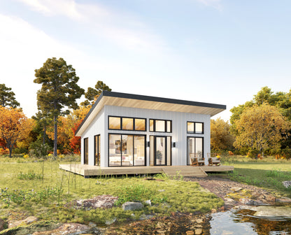A modern single-story cabin with white siding, large black-framed windows, and a wooden deck, shown in a grassy outdoor setting with trees and a small stream under a blue sky.